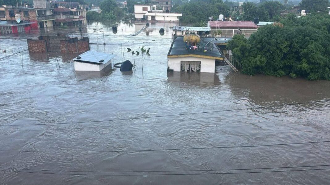 Vista aérea de una zona urbana de Poza Rica, Veracruz, completamente inundada. El agua cubre los cimientos y gran parte de las fachadas de las viviendas, dejando solo visibles los techos. Se observa a tres personas de pie sobre el tejado de una casa blanca, señalando el nivel del agua, presumiblemente a la espera de ser rescatadas.
