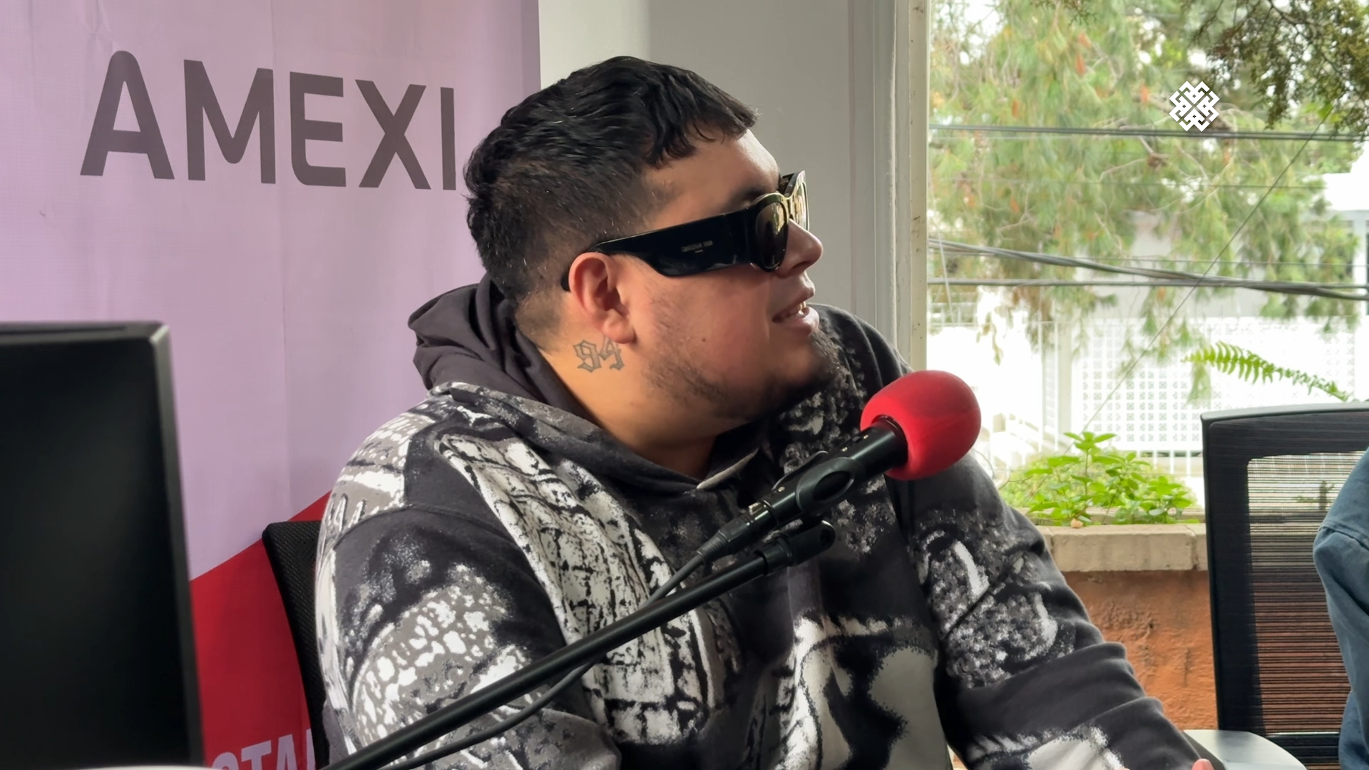 Primer plano de Loojan, integrante de Cachirula y Loojan, usando gafas de sol y hablando en un micrófono rojo durante la entrevista sobre 'Sexolandia'.