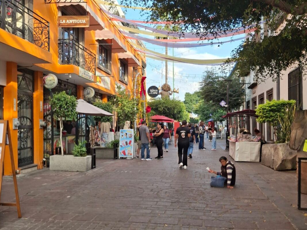Calle peatonal empedrada en el Centro Histórico de Tlaquepaque con edificios coloniales color naranja y decoraciones de papel picado colgando, con personas caminando tranquilamente.