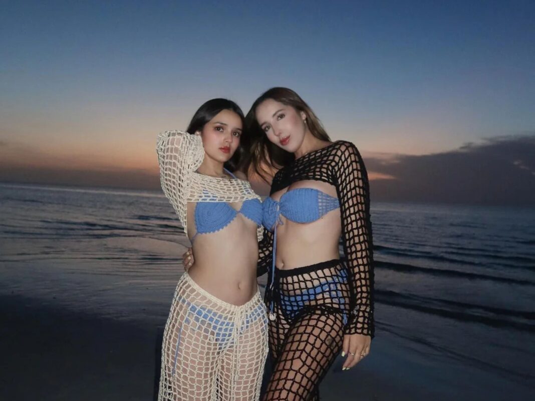Beth Oliver y Kristal Torres posando de pie en la playa al atardecer vistiendo conjuntos de crochet sobre bikinis azules.