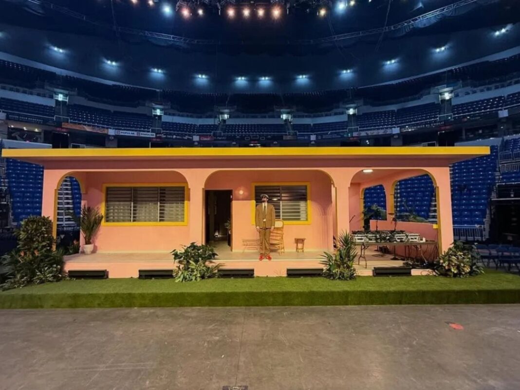 Escenario elevado con forma de casa color rosa y amarillo con techo plano, situado en el centro de una cancha de estadio rodeada de gradas azules vacías. Un hombre con traje café está parado frente a la puerta de la casa.