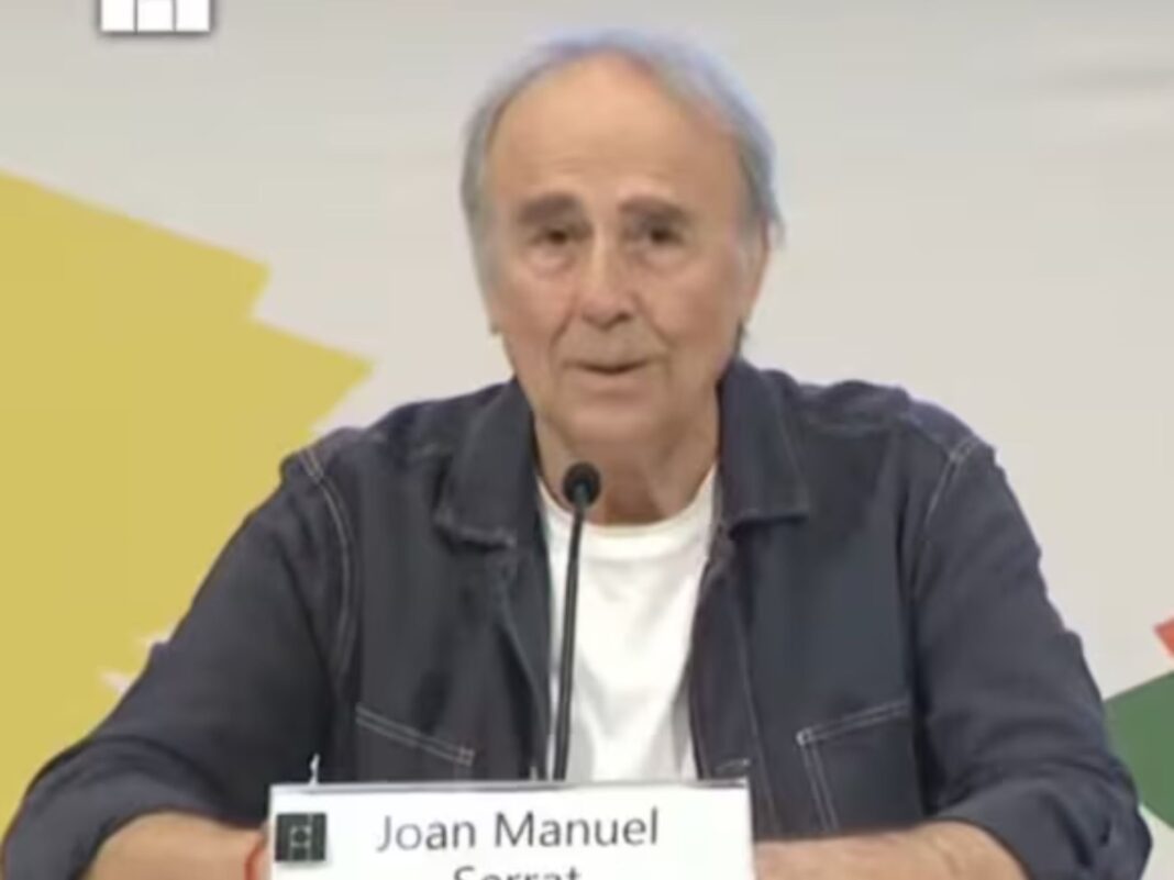oan Manuel Serrat sentado frente a un micrófono durante su charla en la FIL Guadalajara, vistiendo una chaqueta de mezclilla oscura y camiseta blanca, con expresión seria