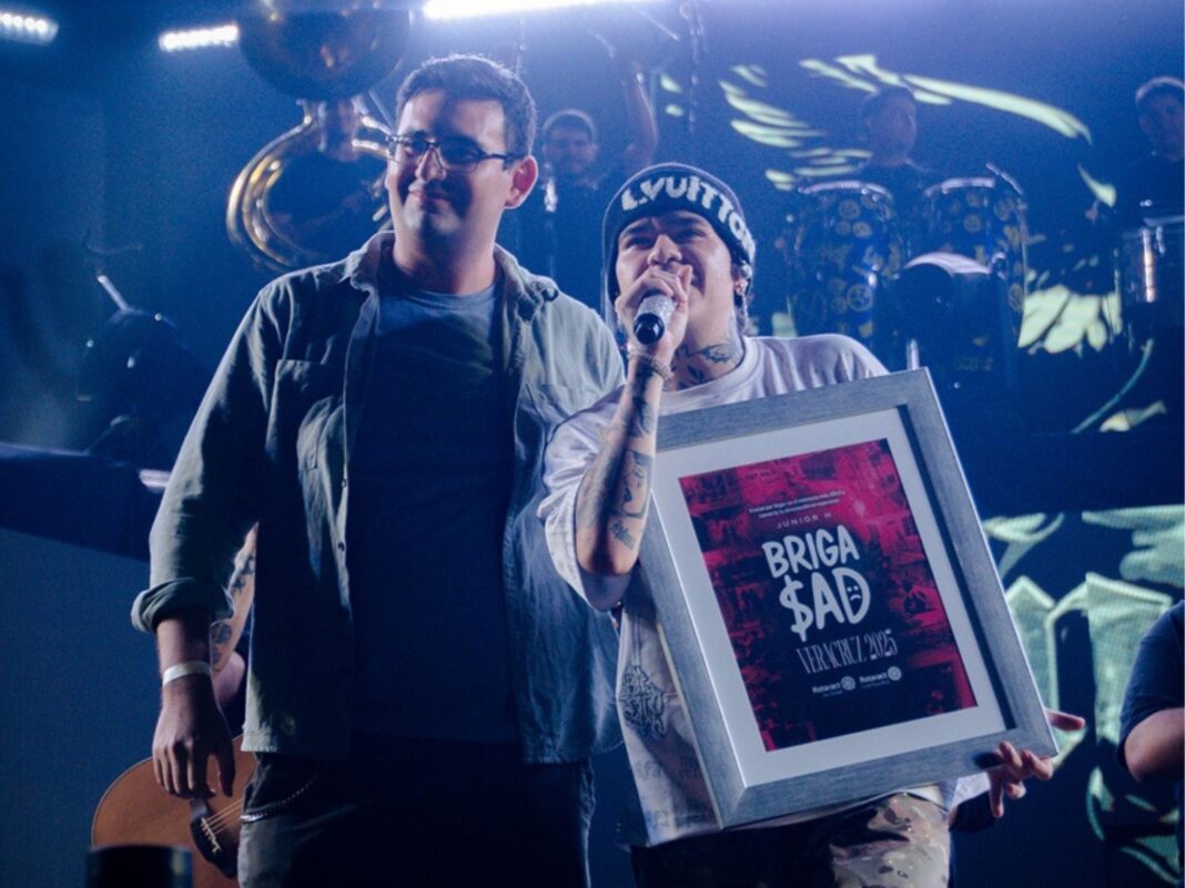 Junior H sostiene un reconocimiento enmarcado con el logo de BrigaSAD junto a un representante de Rotary en el escenario durante su concierto en Veracruz.