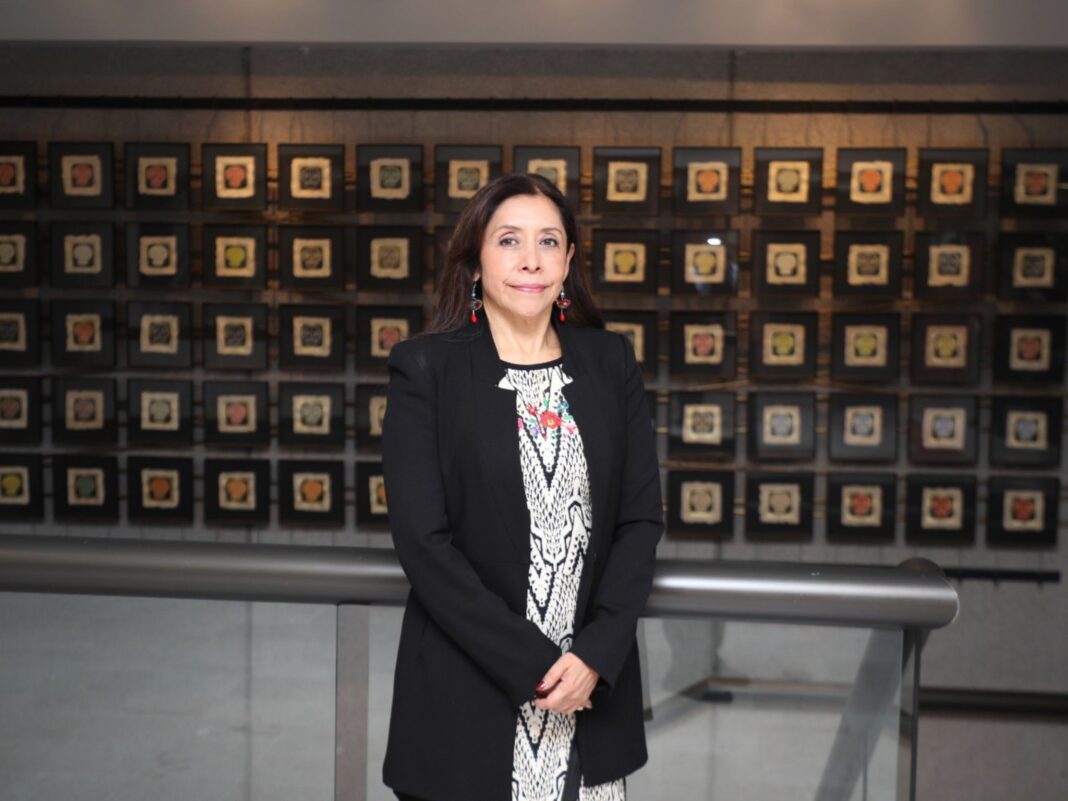 Doctora María Angélica Buendía Espinosa de pie, sonriendo levemente, vistiendo un saco negro y blusa con patrones en blanco y negro, frente a una pared decorada con múltiples cuadros de arte popular mexicano enmarcados.